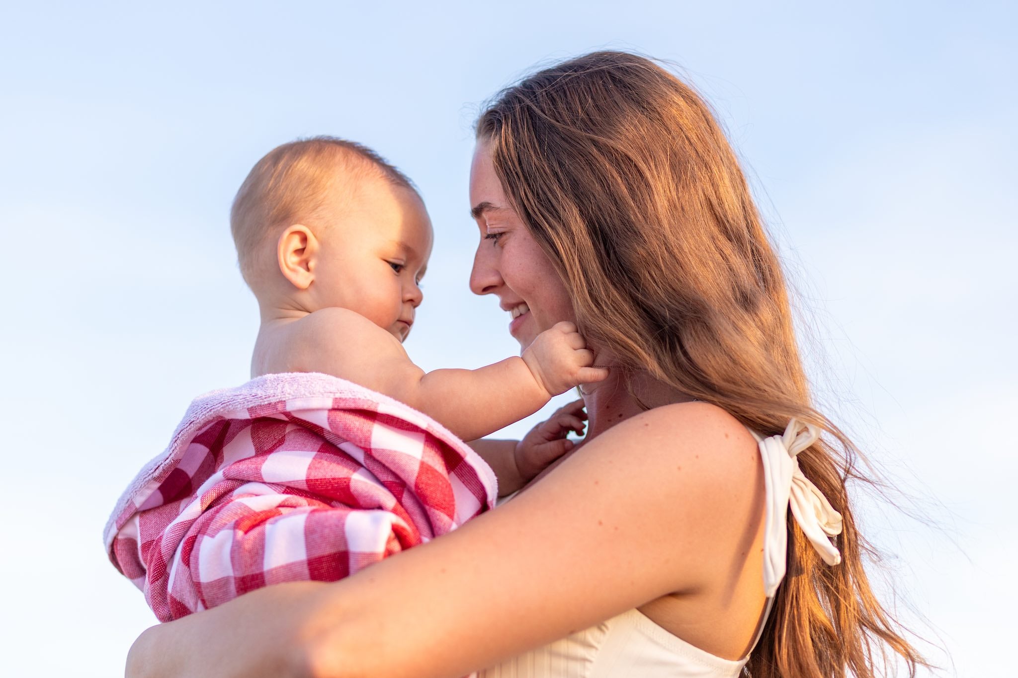 Woman holding a baby wrapped in a red and white Banner gingham red and white towel against a clear blue sky.